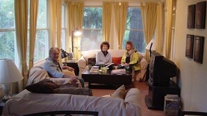 A cozy living room with a sofa set, a coffee table, and a lamp. There are curtains on the windows, and a small table with books and papers.