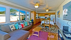 A spacious living area with a sofa, coffee table, and dining table. There is a kitchen area with wooden cabinets and a refrigerator. Large windows provide a view of the outside.