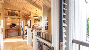 Un intérieur spacieux avec un plafond haut, des poutres en bois, un coin repas avec une table et des chaises en bois, et un escalier doté d’une rampe.