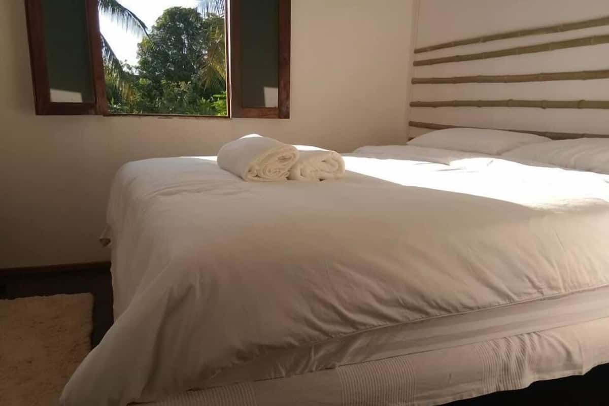 A neatly made bed with white linens and a rolled towel on top, a window with a view of greenery, and a bamboo headboard.
