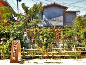 Une maison traditionnelle japonaise, avec une façade en bois, entourée d’une végétation luxuriante et d’une clôture en bambou.