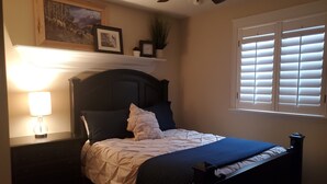 A bedroom with a bed, a nightstand, a lamp, a ceiling fan, a window with blinds, and wall-mounted shelves with framed pictures and a plant. - Great Sandy apartment with hot tub and pool table! (Sandy)