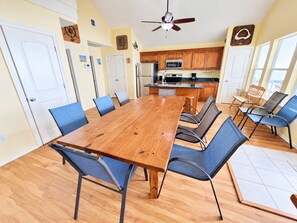 A dining area with a wooden table and chairs, a kitchen in the background, and a ceiling fan. - Wine Down: Coastal Realty Group (Grand Isle)