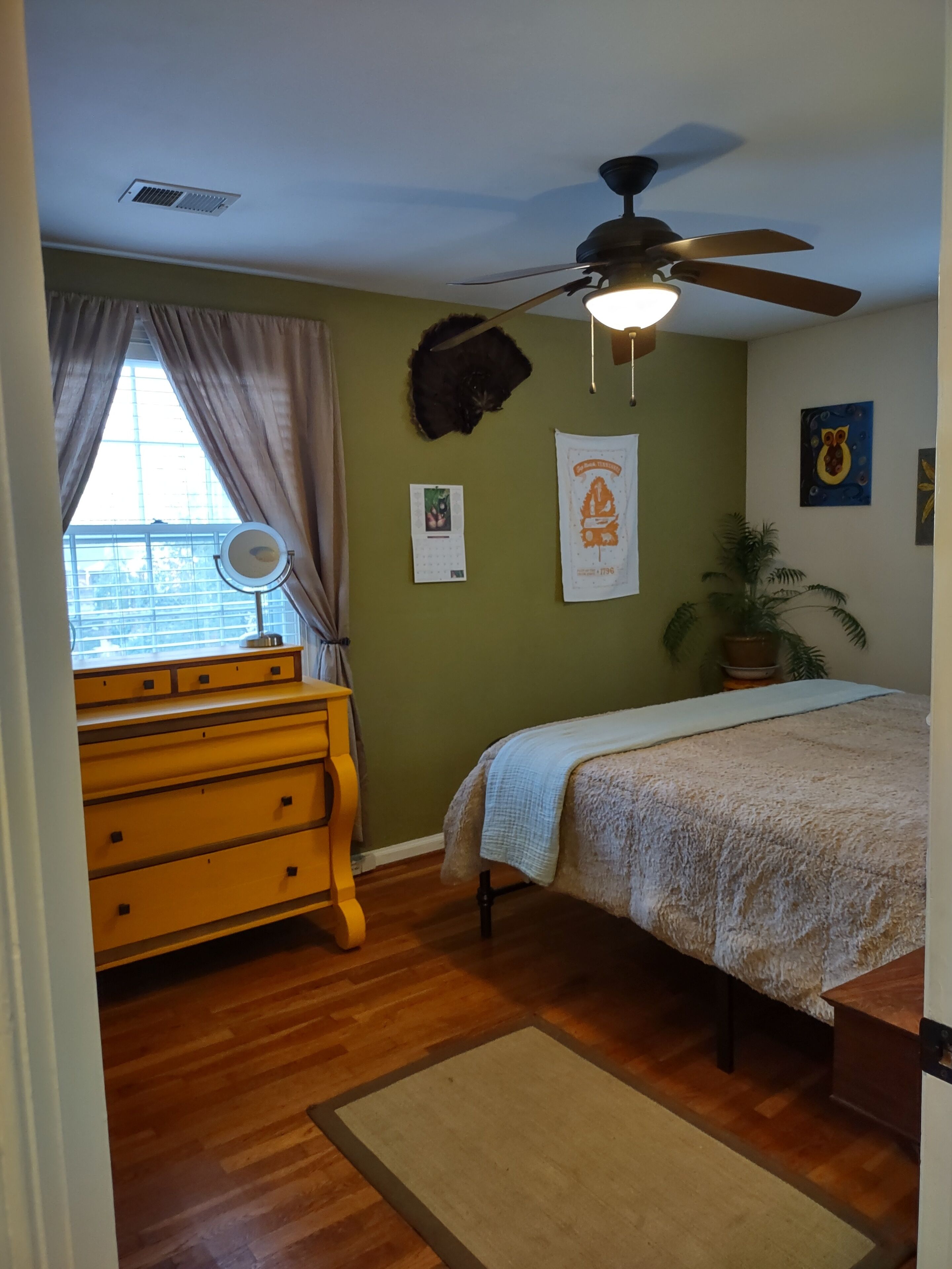 Quarto com cama, cômoda, ventoinha no teto, janela com cortinas e parede com quadros.