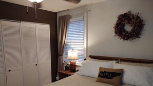 A bedroom with a bed, a nightstand with a lamp, a window with blinds, a wardrobe, and a wall decoration.