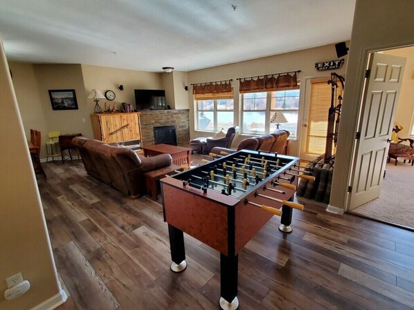 A cozy living room with a foosball table, comfortable seating, a fireplace, and a view of the outdoors.