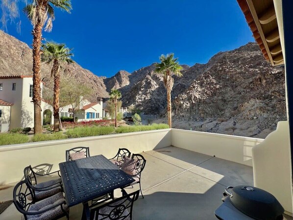 An outdoor patio with a table and chairs, a grill, and a view of a mountainous landscape with palm trees.