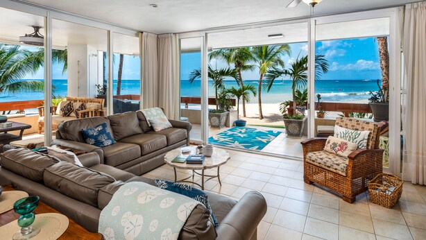 A spacious living area with a large sofa, a wicker chair, and a small table. There is a view of the beach through large glass doors. - Three Steps Onto the Most Beautiful Beach in Cozumel (cozumel)
