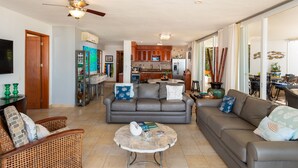 A spacious living area with a grey sofa, a wicker chair, and a coffee table. A kitchen with wooden cabinets and a dining area with a table and chairs are visible in the background. - Three Steps Onto the Most Beautiful Beach in Cozumel (cozumel)