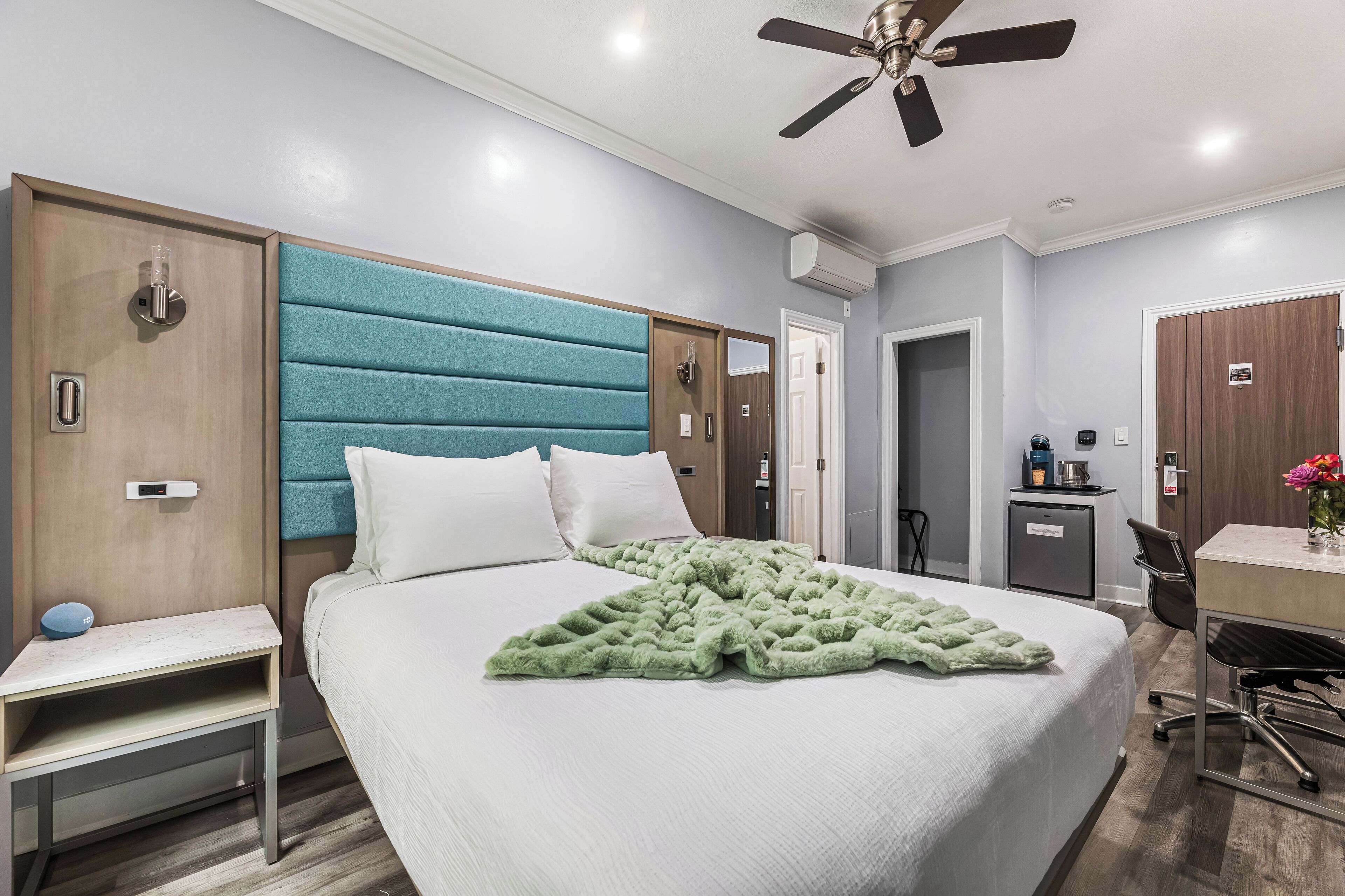 Une chambre d’hôtel équipée d’un lit, d’un bureau, d’une chaise, d’un ventilateur de plafond et d’une petite kitchenette.