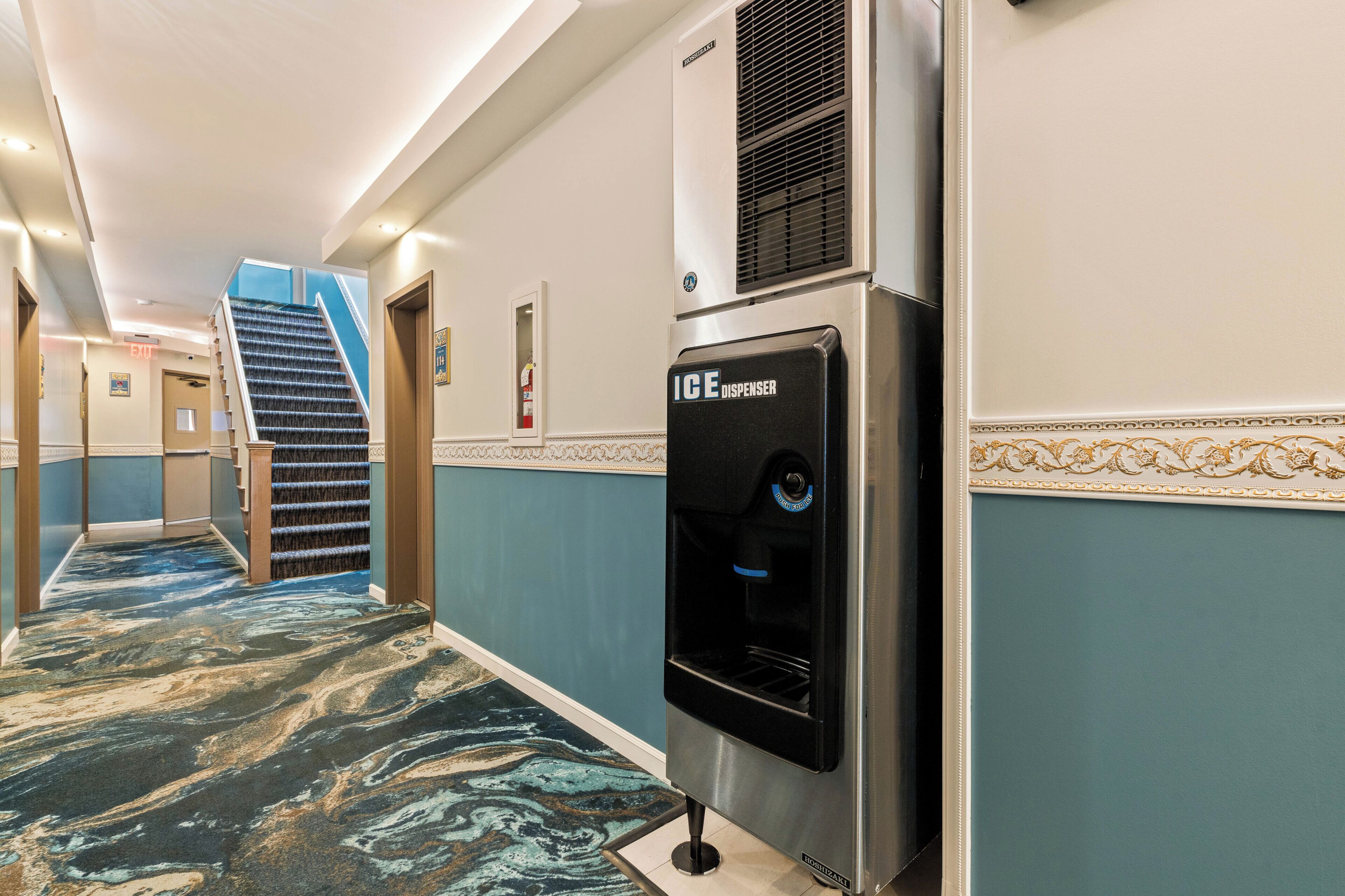 Corridoio di un hotel con tappeto a motivi marmorizzati, un dispenser d'acqua, una scala e una porta con il simbolo del numero.