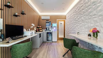 Una moderna zona de recepción con un panel de madera, mostrador de recepción, refrigerador, microondas y una pequeña sala de estar con sillas verdes.