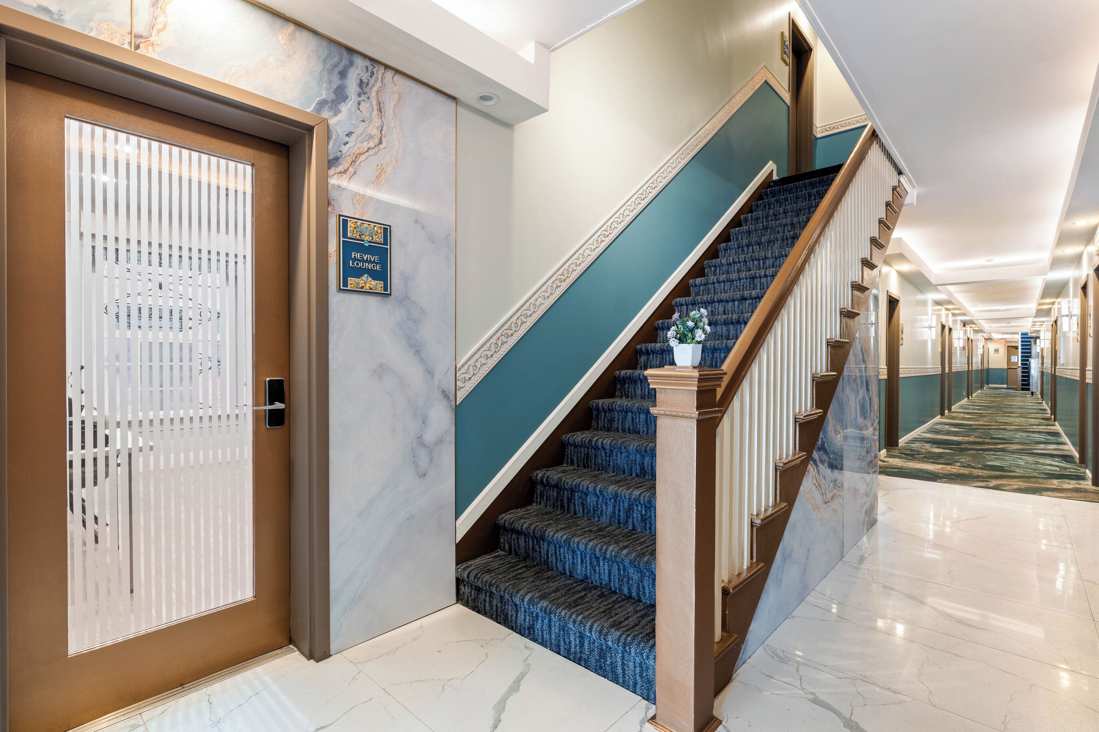 Un vestíbulo de hotel con piso de mármol, una escalera con peldaños cubiertos de moqueta azul y una puerta con persianas verticales.