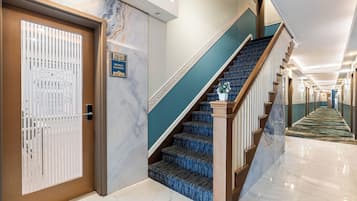 Un vestíbulo de hotel con piso de mármol, una escalera con peldaños cubiertos de moqueta azul y una puerta con persianas verticales.