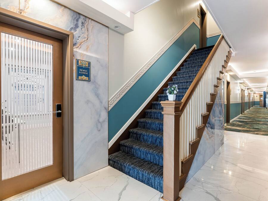 Un vestíbulo de hotel con piso de mármol, una escalera con peldaños cubiertos de moqueta azul y una puerta con persianas verticales.