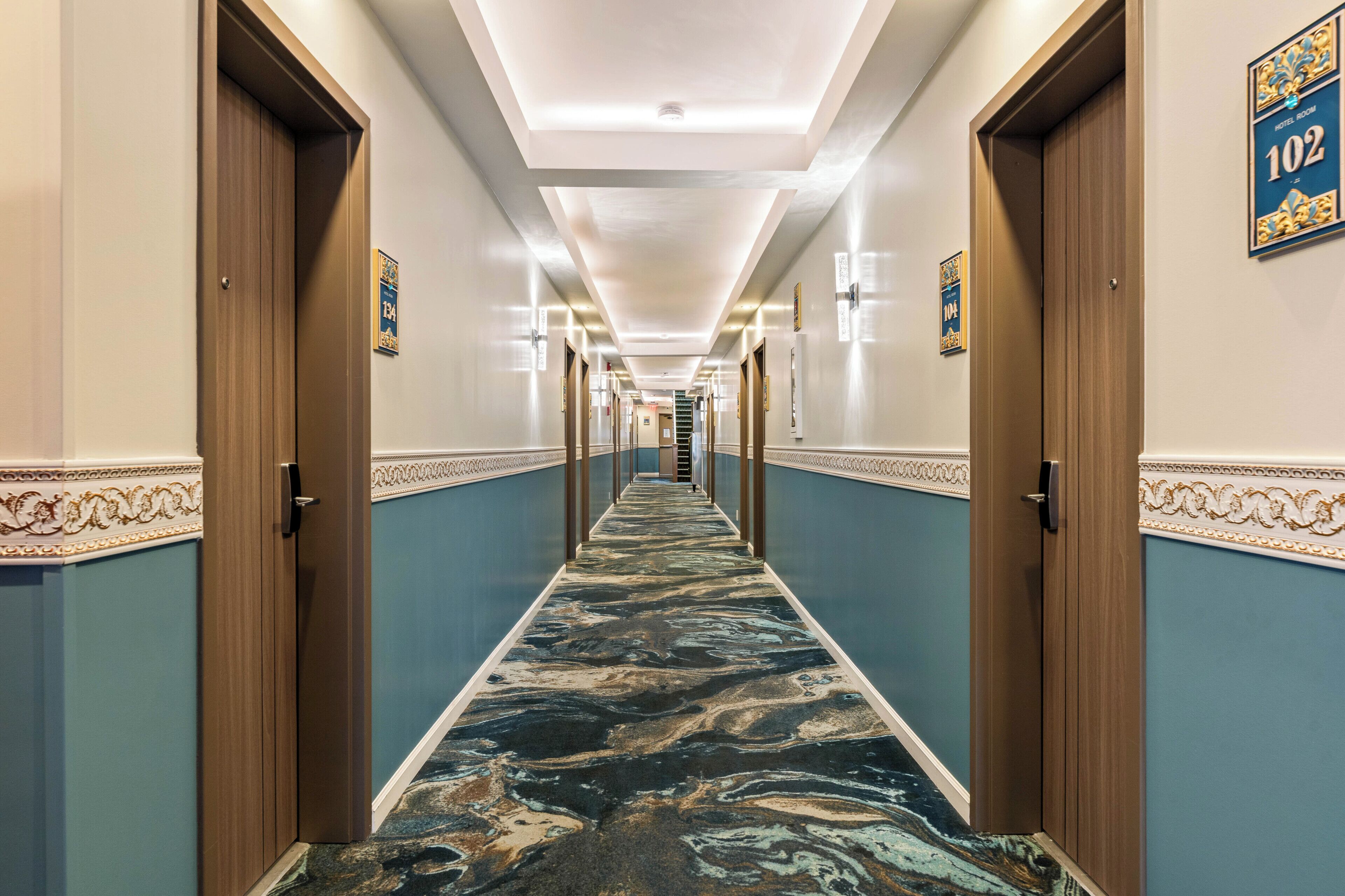 A hotel hallway with doors numbered 102 and 104, a marble-patterned carpet, and a ceiling with recessed lighting.