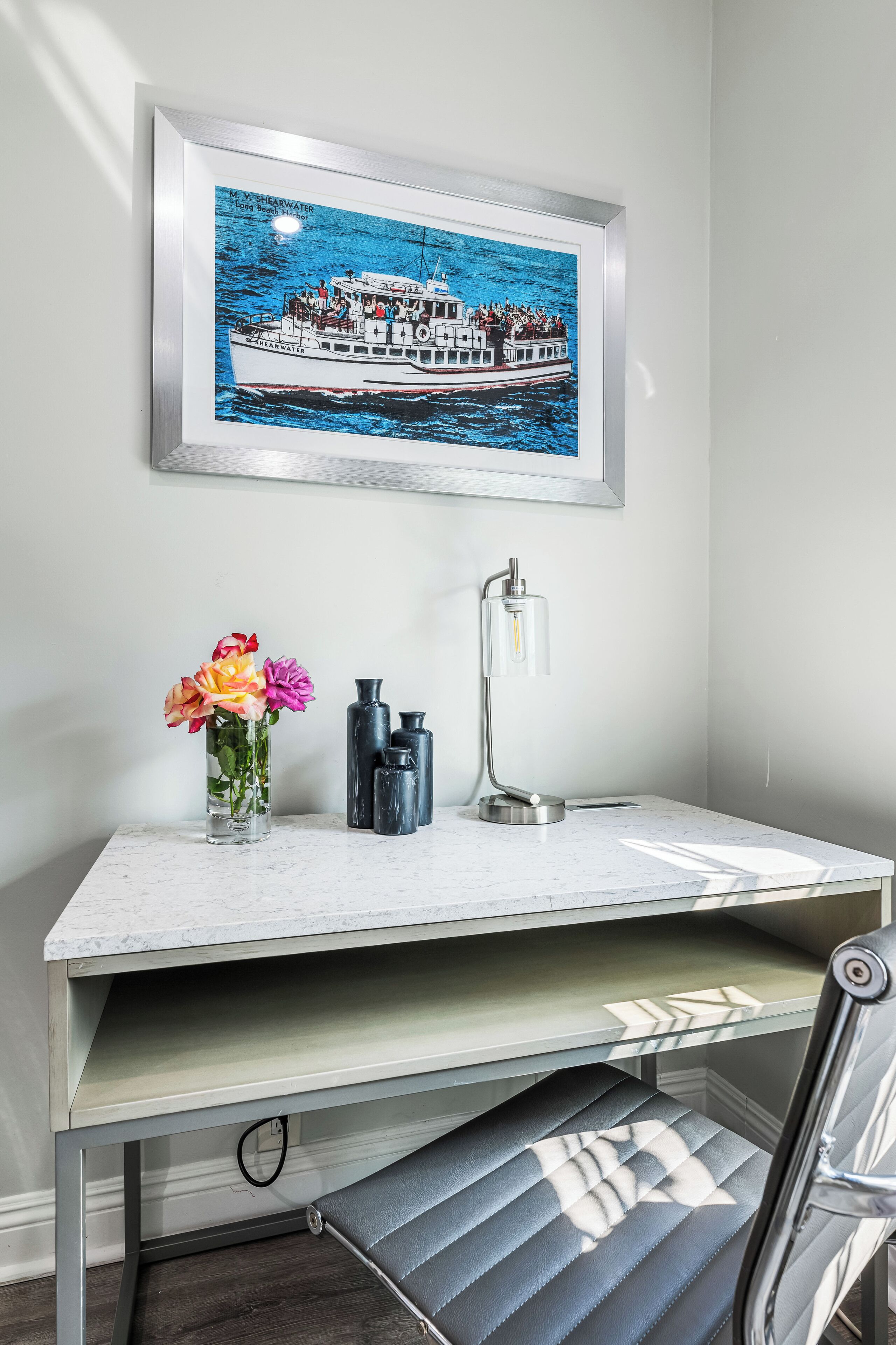 A desk with a framed picture of a boat, a vase with flowers, and a desk lamp.