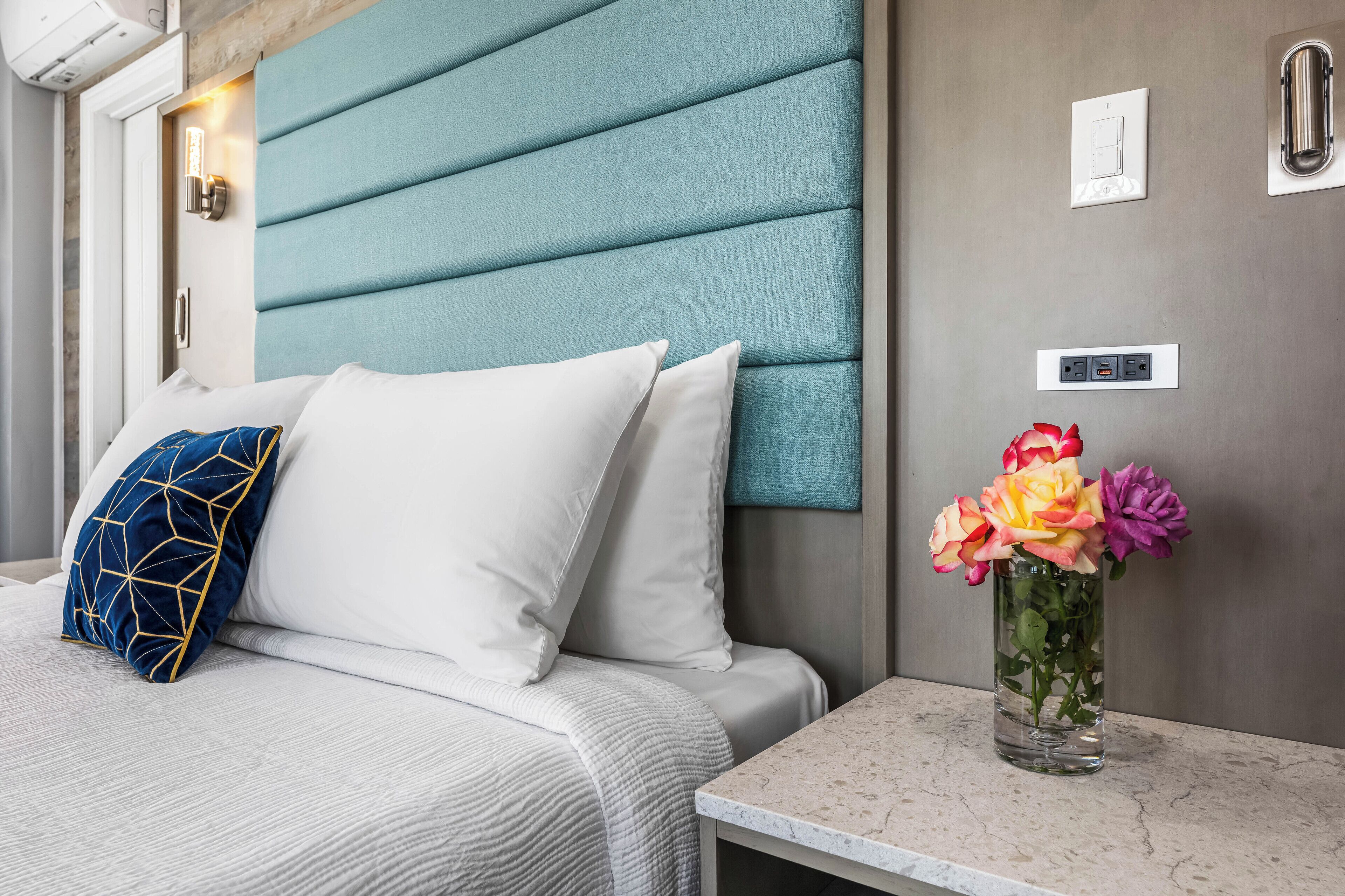 A neatly made bed with white and blue pillows, a vase with flowers, and a marble bedside table.