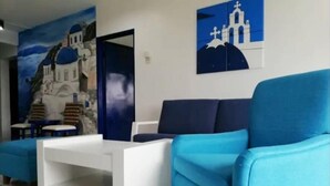 Ein modernes Wohnzimmer mit einem blauen Sofa, einem weißen Couchtisch und einem Wandbild einer griechischen Insel.