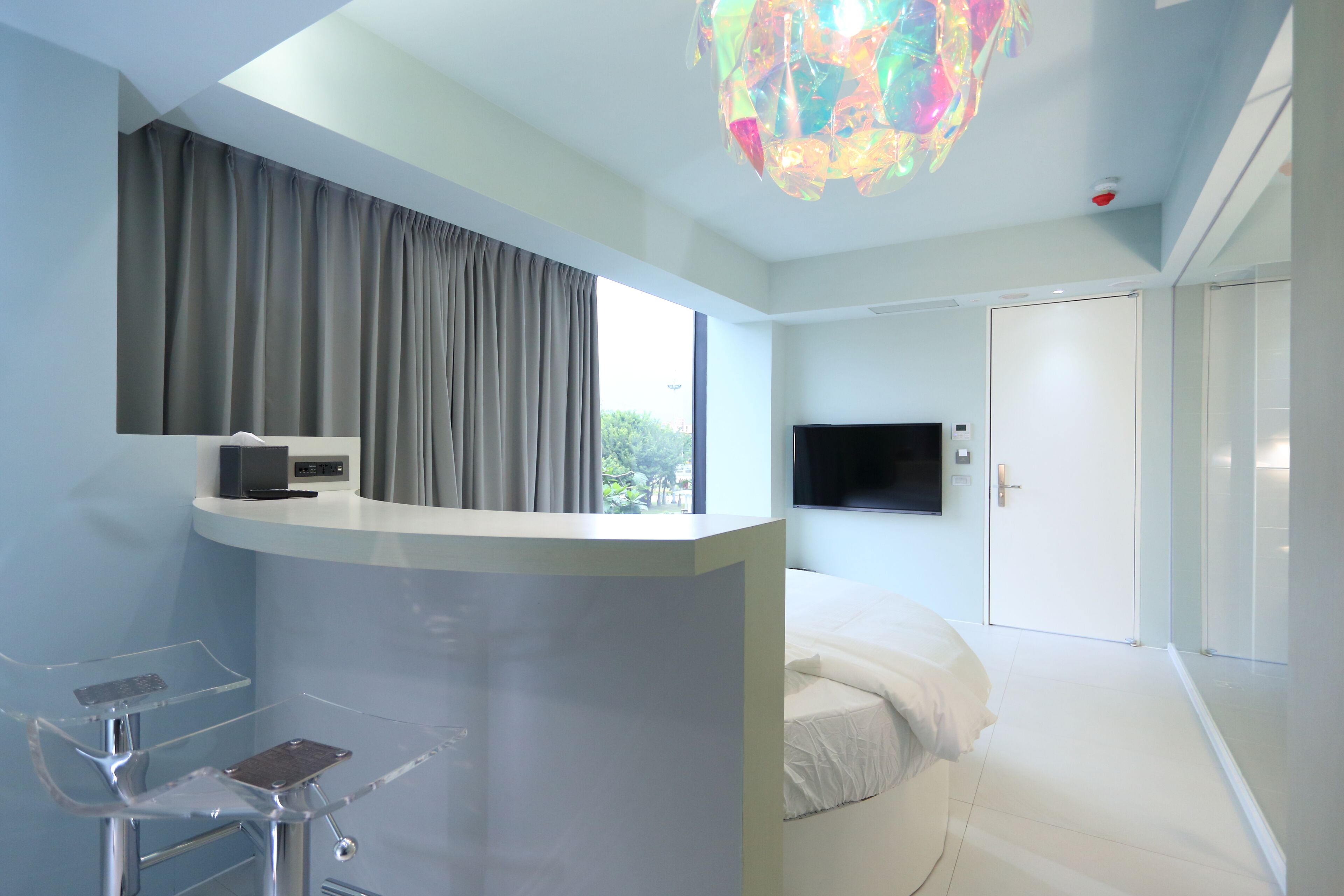 Un dormitorio moderno con cama, escritorio, televisor y un amplio ventanal con cortinas.