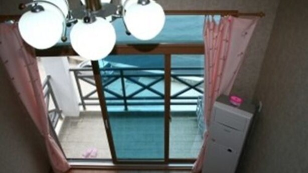 Ein Zimmer mit einer Schiebetür und einem Balkon mit Meerblick. Über der Tür hängt eine Deckenleuchte und rechts daneben befindet sich ein kleines Schränkchen.