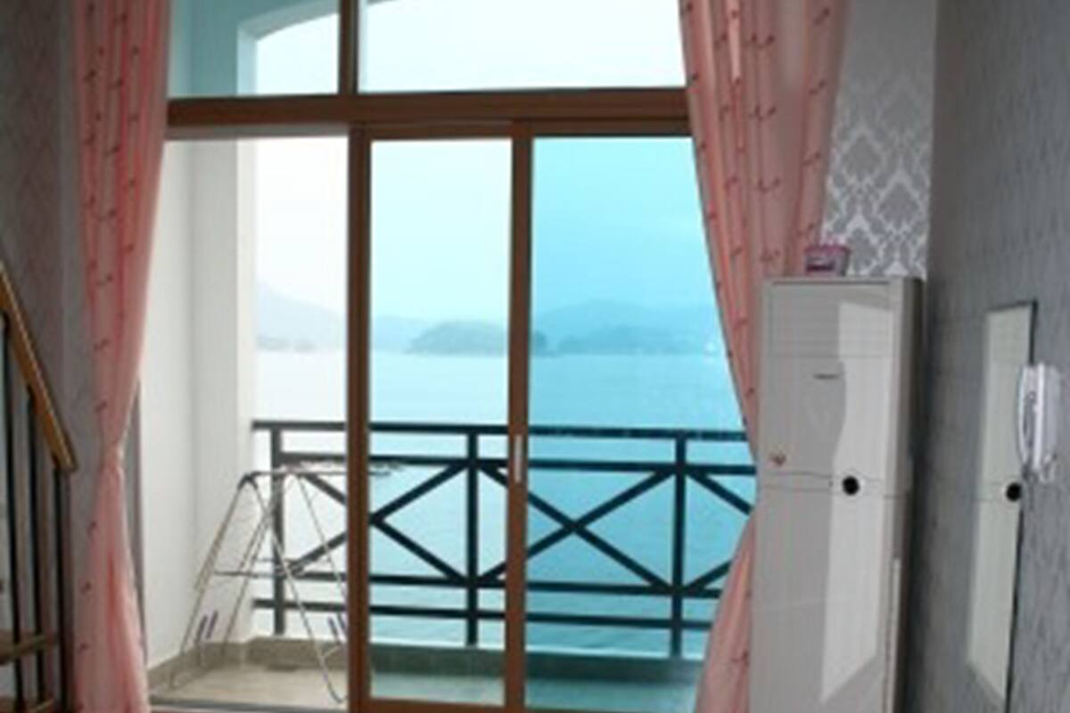 Une pièce avec un plancher en bois, un réfrigérateur blanc et une vue sur la mer, visible à travers une grande fenêtre ornée de rideaux roses.