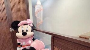 A plush Minnie Mouse sitting on a wooden bench in a room with a wooden door and a hanging light fixture. - Chamie B&B (Hengchun)