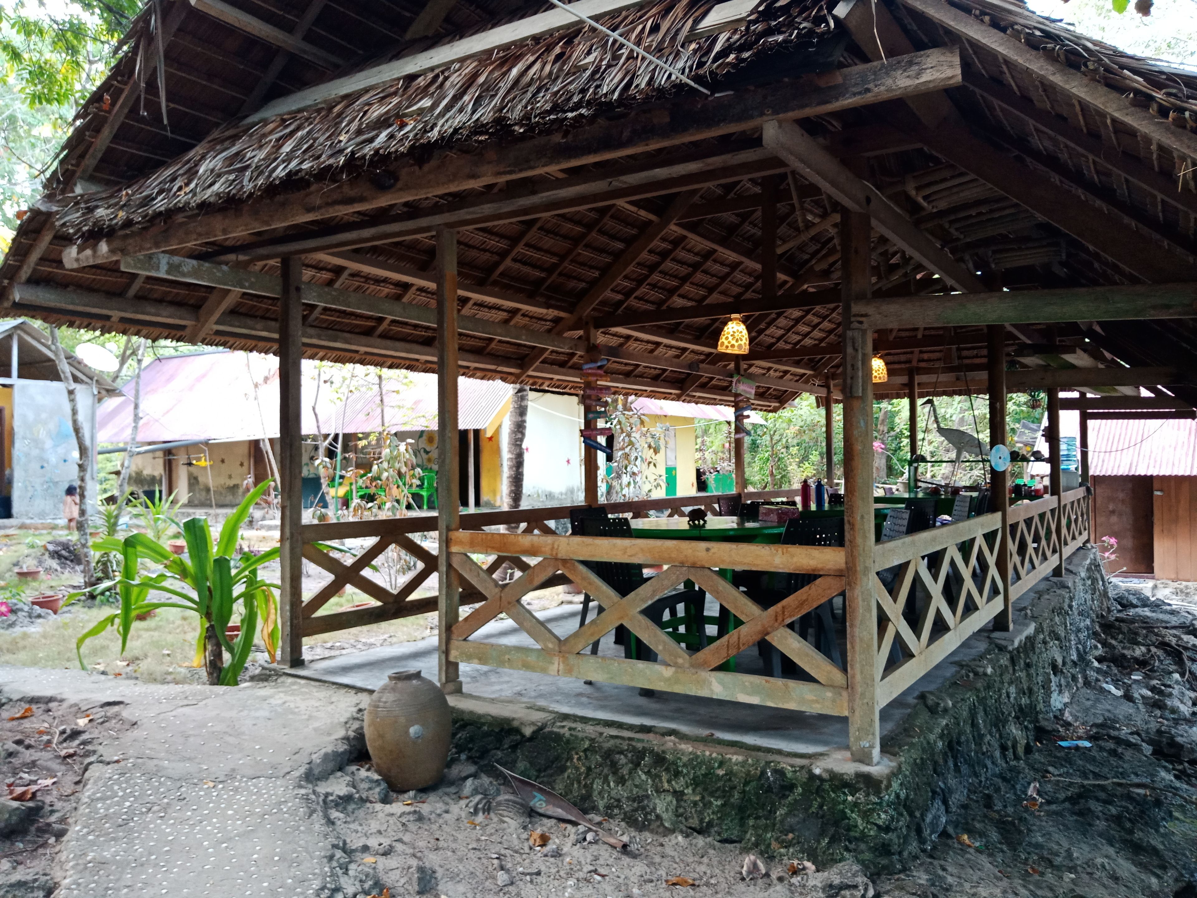 A rustic wooden pavilion with a thatched roof, surrounded by greenery and a paved area.