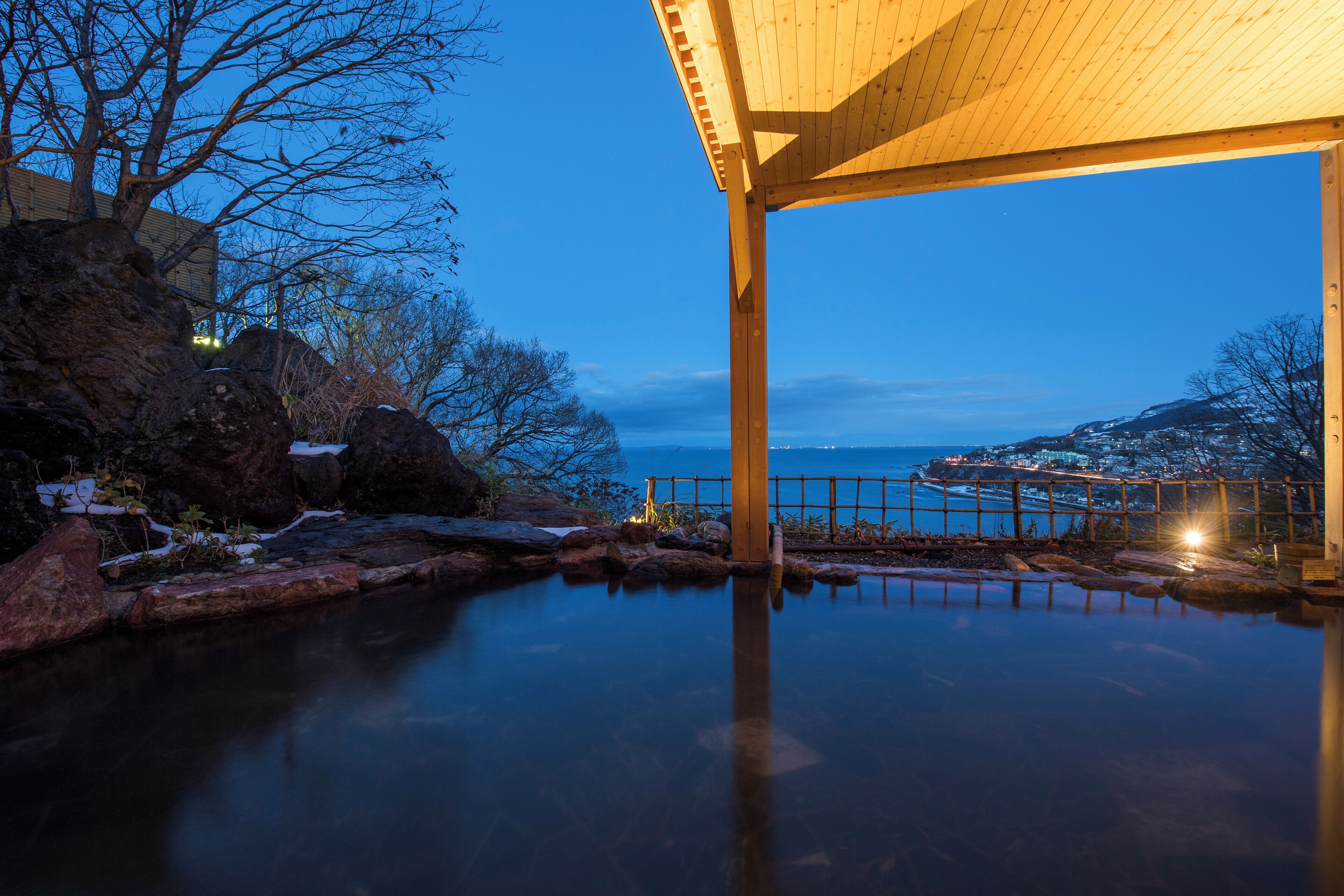 A tranquil outdoor pool area with a wooden pergola, surrounded by rocks and snow patches, overlooking a coastal town.