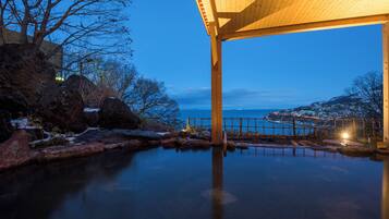 A tranquil outdoor pool area with a wooden pergola, surrounded by rocks and snow patches, overlooking a coastal town.