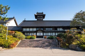 Un edificio japonés tradicional con techo de tejas, una escalera de madera que conduce a la entrada y un cielo azul despejado.