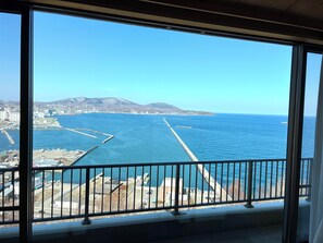 Vista desde un balcón con barandilla, sobre un cuerpo de agua con muelle y una costa lejana con edificios y colinas.
