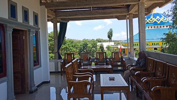 A covered outdoor seating area with wooden furniture and a view of a colorful building in the background. - Wakatobi Dive Inn (Kaledupa Island)