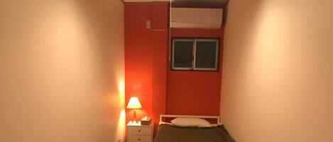 A narrow room with a bed, a desk, a chair, a nightstand, a lamp, and a ceiling fan.