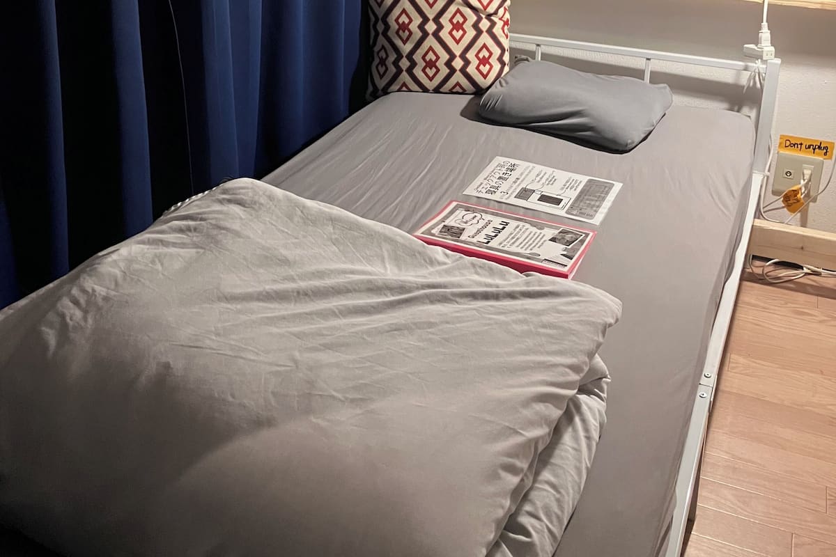 A single bed with a grey mattress, a white pillow, a patterned pillow, and a magazine on the bed.