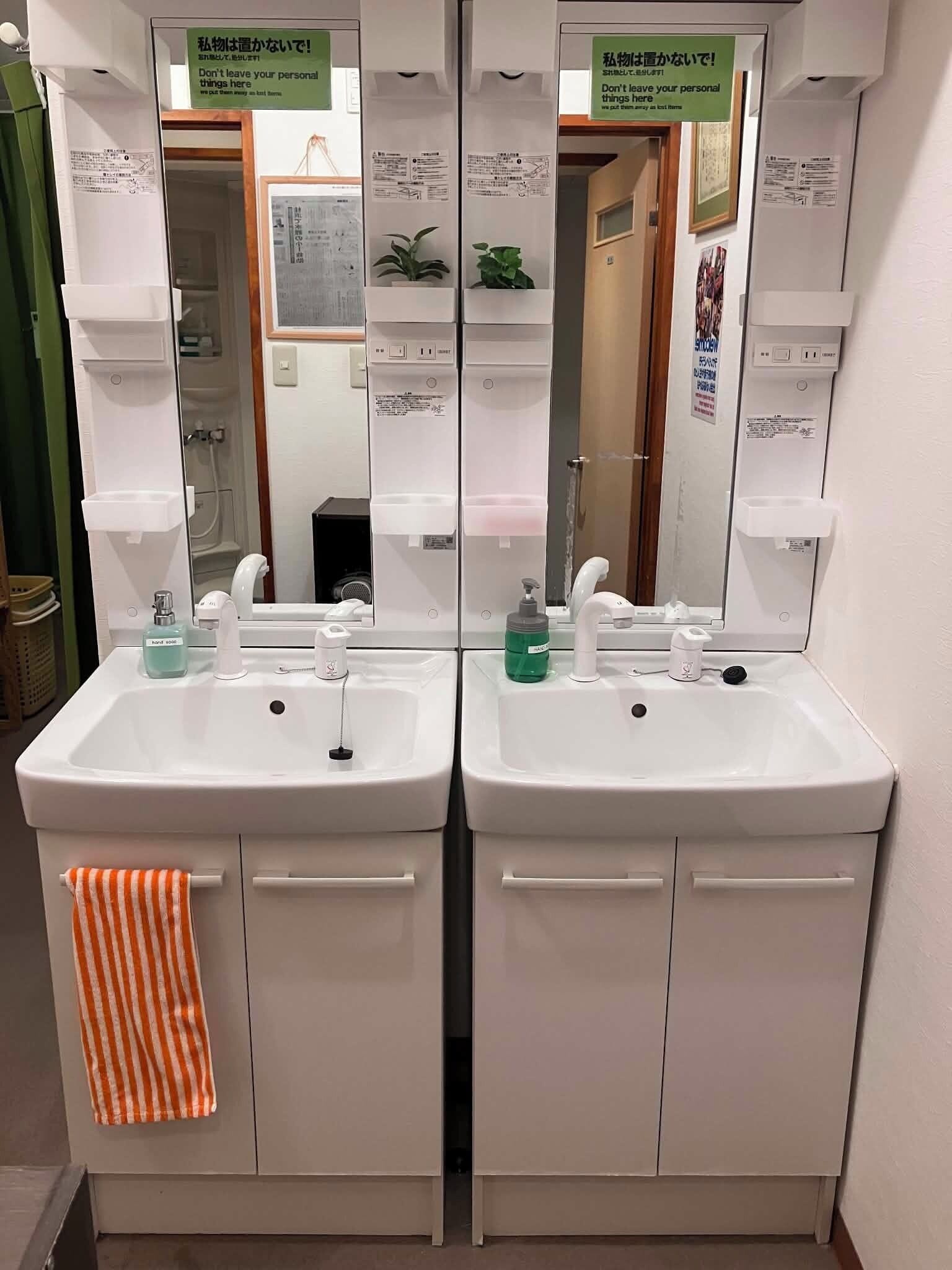 A bathroom with two sinks, each with a mirror above. There are hand soap dispensers and a towel rack with an orange towel.