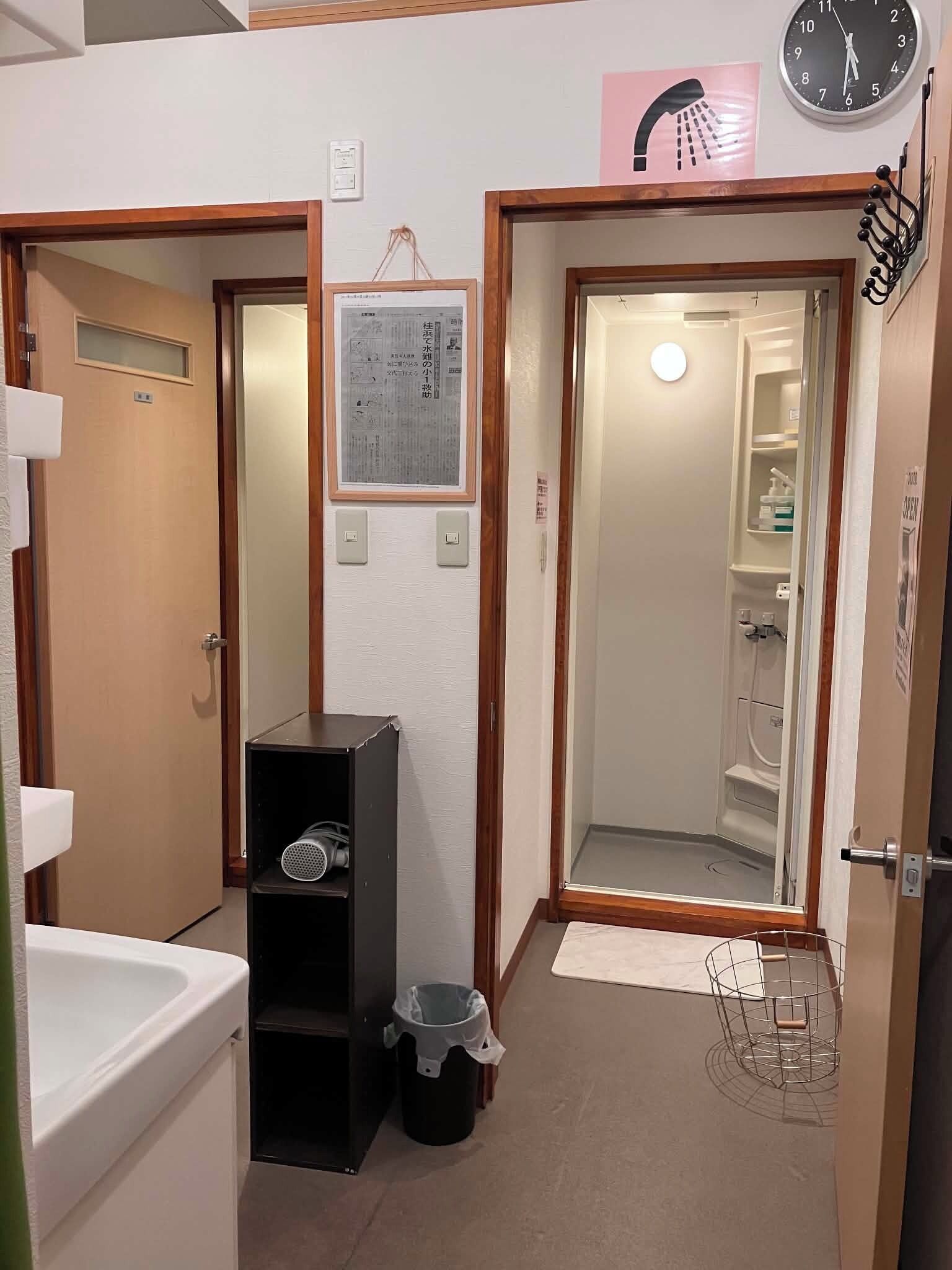 Un baño pequeño con lavamanos, un cubo de basura, zona de ducha y un reloj en la pared.