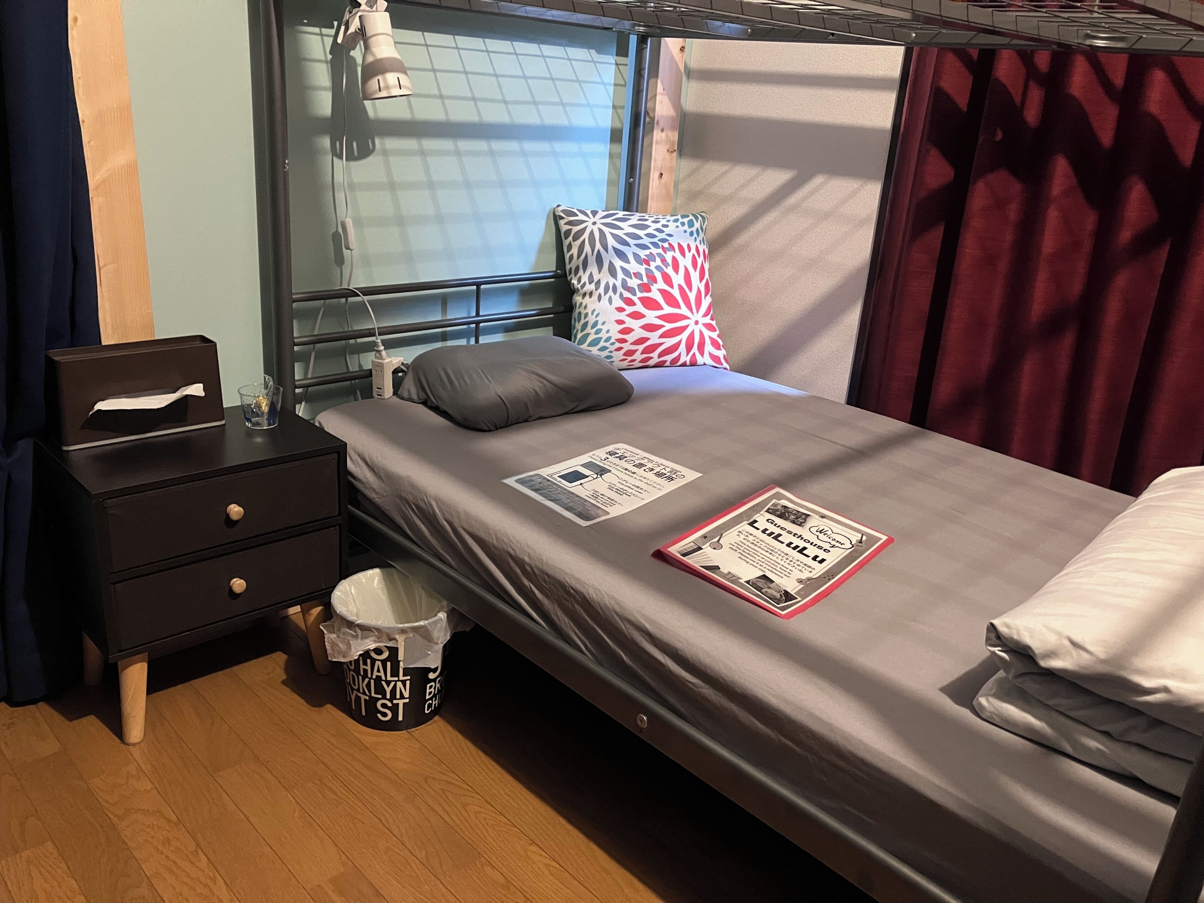 A single bed with a grey mattress, a pillow, a bedside table with a tissue box, a lamp, and a bucket with a label.