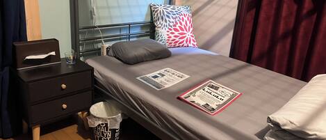 A single bed with a grey mattress, a pillow, a bedside table with a tissue box, a lamp, and a bucket with a label.