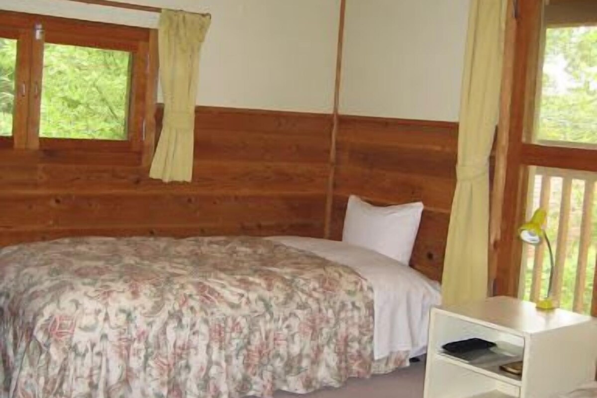 A room with two beds, wooden beams on the ceiling, a window with curtains, and a bedside table with a lamp.