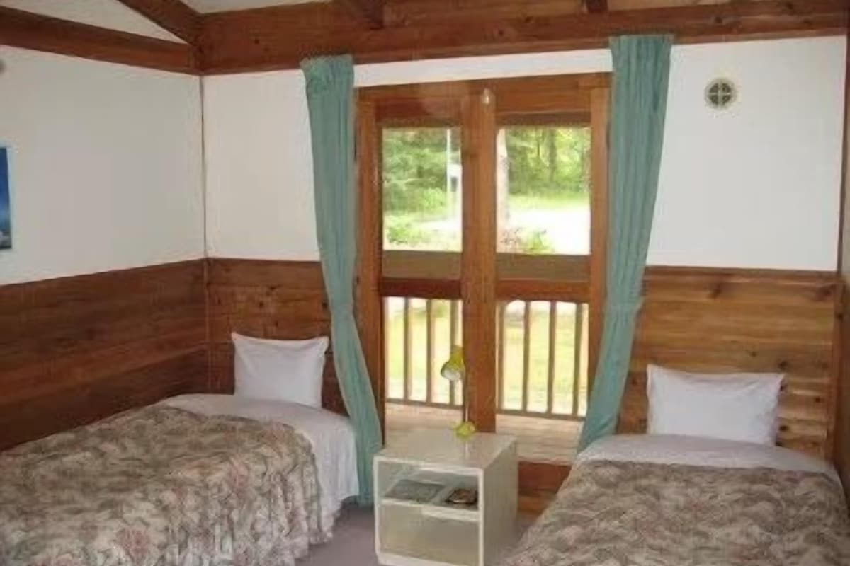 A room with two beds, wooden walls, a window with curtains, and a small table.