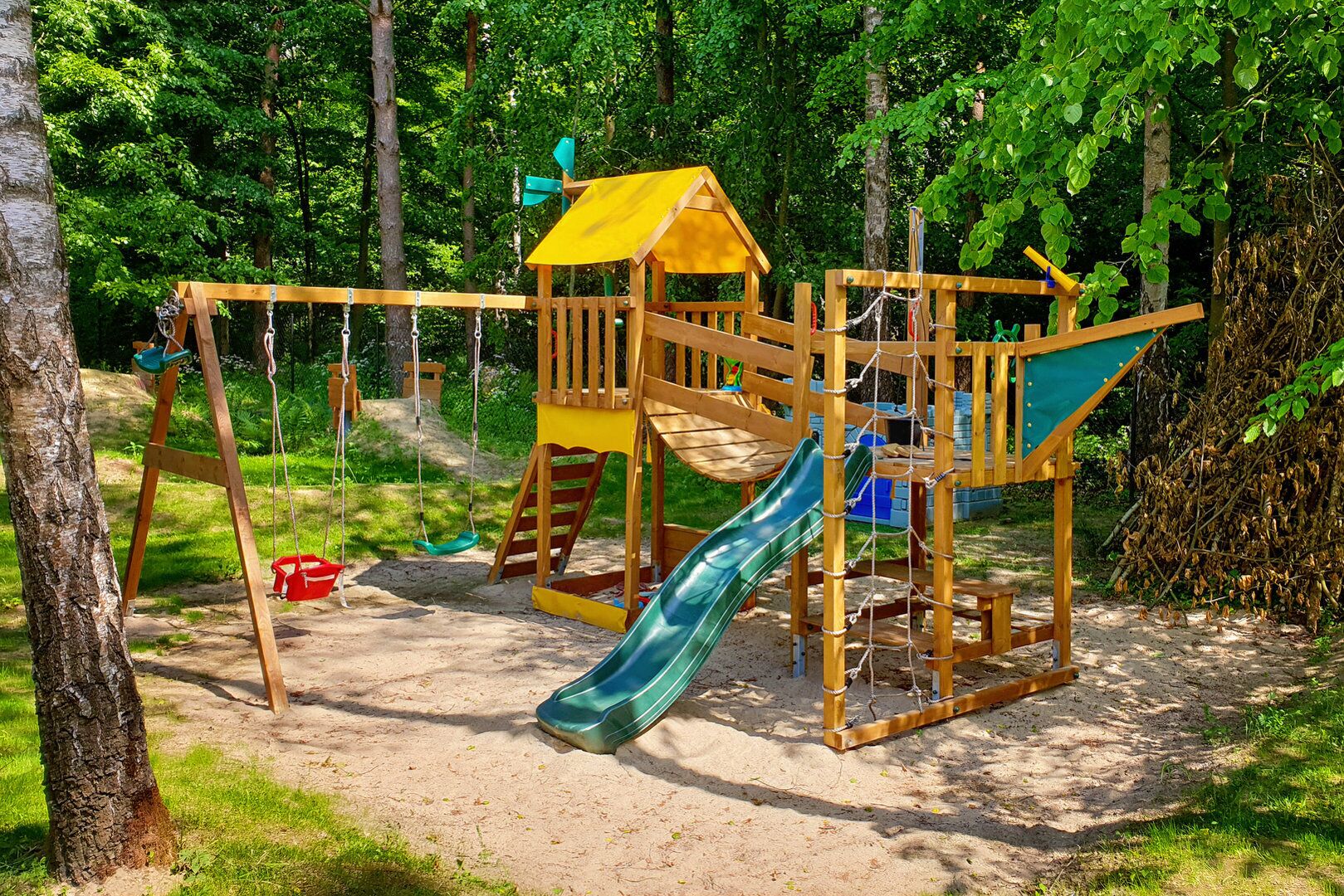 Un parc infantil de fusta amb tobogan i cadira de balanceig, envoltat d'arbres.