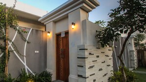 A modern house with a wooden door, a white wall with a geometric pattern, and a small garden with plants and a tree. - Giri Ungasan Villa (Ungasan)
