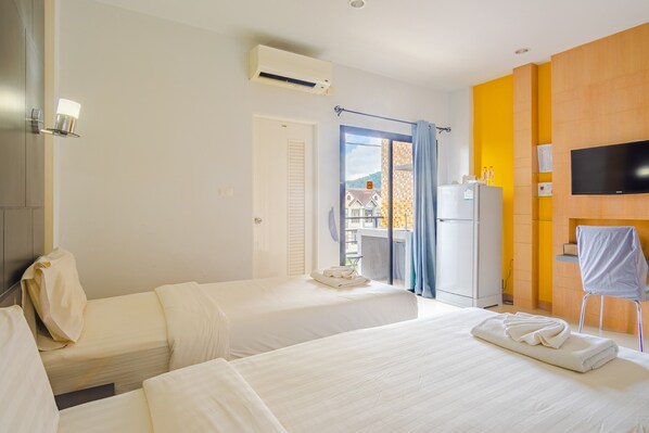 A hotel room with two beds, a TV, an air conditioning unit, and a view of a building through a glass door. - Phoomjai House (Chalong)