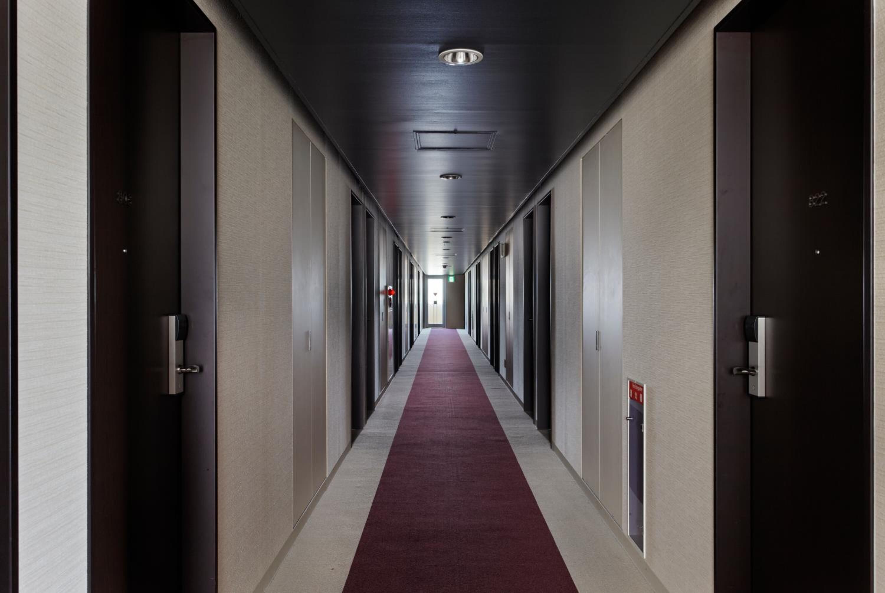 A long, narrow hallway with a red carpet, doors on both sides, and a ceiling with recessed lighting.