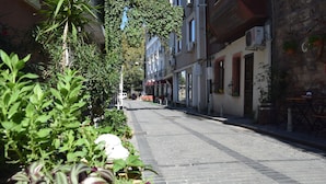 Une ruelle étroite pavée, bordée d’immeubles, dont certains sont dotés de balcons et de plantes en pot.