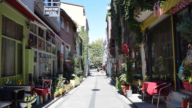 Une rue étroite bordée d’immeubles, dont l’hôtel Palace Point. On y trouve des espaces de restauration en plein air avec des tables et des chaises, et des plantes en pot sont disposées le long des trottoirs.