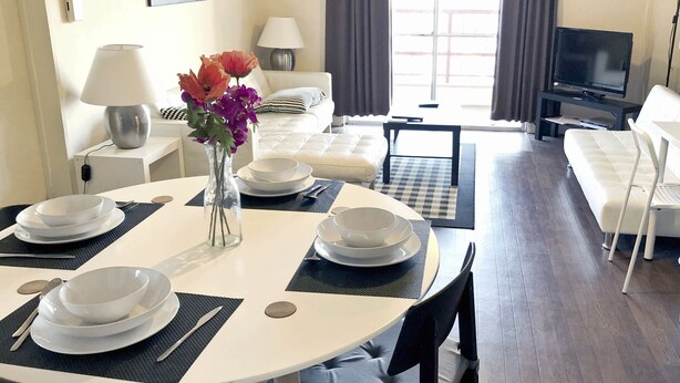 A dining area with a round table set for four, a vase with flowers, and a view of a living room with a sofa and a television. - Spacious Apartment - 5-7 minute walk to Shinkansen and Shin-Kobe Subway Station (Kobe)