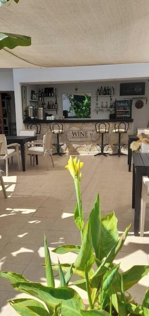 Un área exterior con mesas y sillas, una barra con estantes llenos de botellas y un letrero que dice 'WINE'.