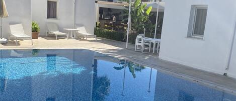 A clear blue pool with tiled flooring, white lounge chairs, and a small table.
